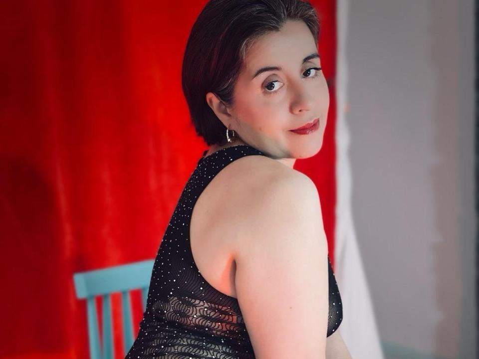 Dalila69s with short brown hair, wearing a black sparkly dress, looking over her shoulder, smiling, showing her bare shoulders and back, sitting on a blue chair.