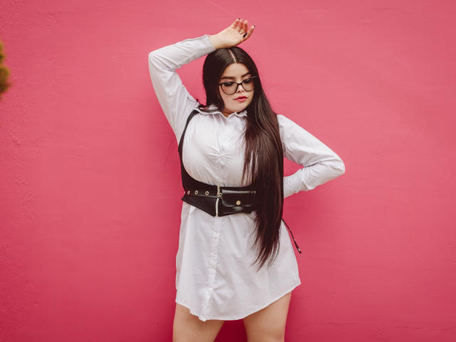 BrithneyLee with long black hair, glasses, white dress, black belt, standing against pink wall, hand on head, hand on hip, showing big tits, cute, sexy.