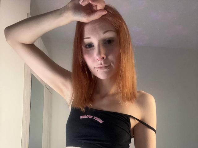 HannaCool with red hair, black strap-down top, small tits, nose ring, and mole above her lip, raising her arm, looking at the camera.
