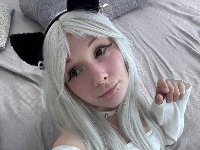 HannaCool with white hair, black cat ears, nose ring, lip piercing, and long eyelashes, wearing a white top, lying on a bed, looking up with a slight smile.