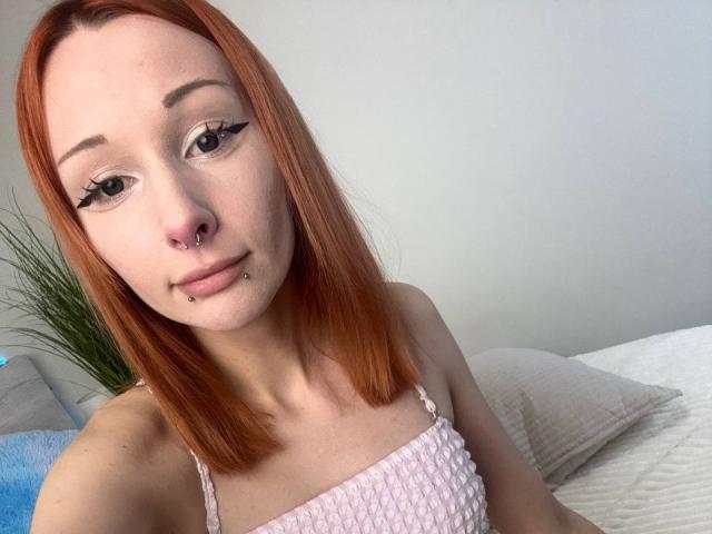 HannaCool with red hair, nose piercing, lip piercing, and eyeliner, wearing a pink, textured top, has a cute, slightly smug expression, and is sitting on a bed.