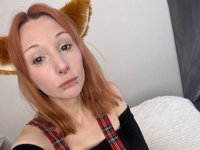 HannaCool has red hair, cat ears, a nose ring, and a lip piercing. She's wearing a red plaid dress over a black top. Her tits are not visible.