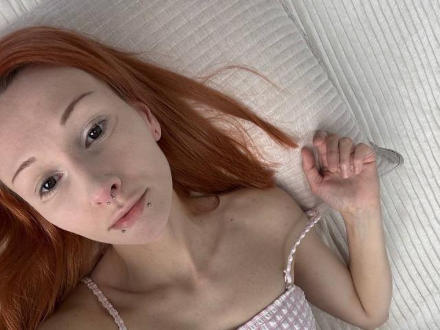 HannaCool with red hair, nose ring, lip piercing, and ear piercings, lying on a white pillow, wearing a pink and white checkered top, holding a silver ring.