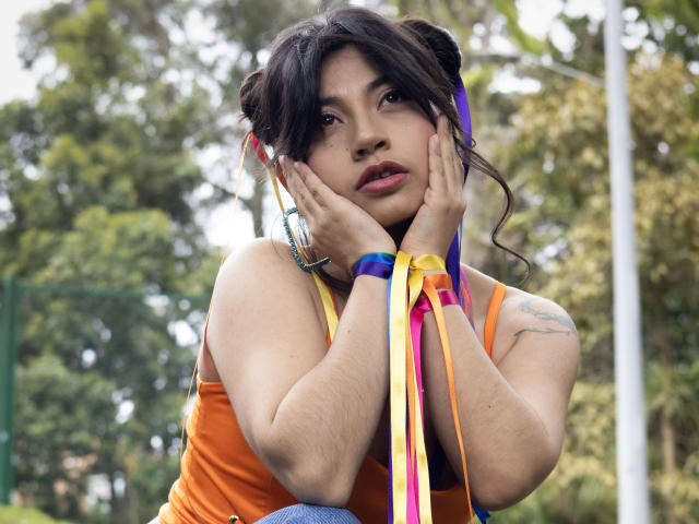 SeleneLeon, mit zwei Duttfrisuren und dunklem Haar, trägt ein orangefarbenes Tanktop, hat die Hände auf den Wangen, bunte Bänder und ein Armband, und wirkt ernst, wobei ein sichtbares Tattoo auf ihrem rechten Arm zu sehen ist..