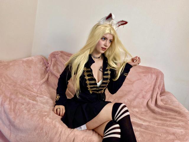 MarriAnni with blonde hair, wearing a black dress with gold trim, white thigh-high socks, and fox ears, sitting on a pink-covered couch, holding her hair.
