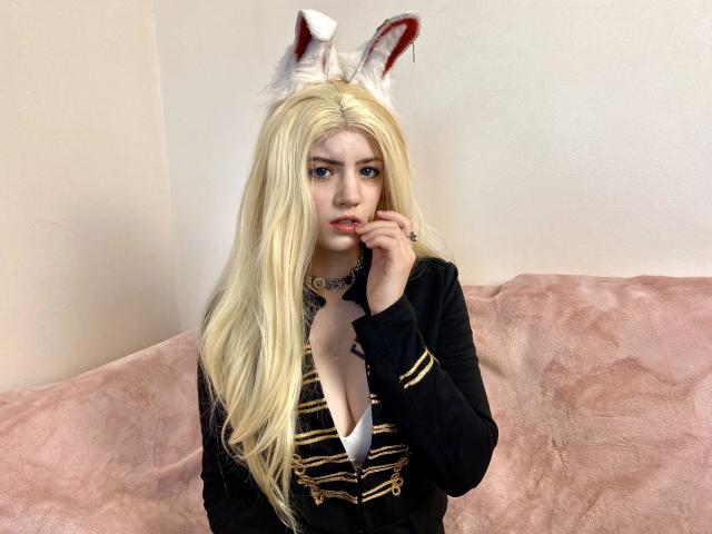 MarriAnni met lang blond haar, witte konijnenoren, een zwarte jas met gouden details, een diepe decolleté, een wit shirt, een roze bank, een hand in de buurt van de mond, die naar de camera kijkt, bleke huid, blauwe ogen..