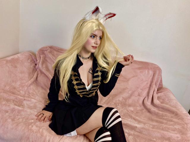 MarriAnni with blonde hair, white bunny ears, black dress with gold trim, exposing her boobs, black skirt, striped thigh-highs, sitting on pink couch, holding hair.