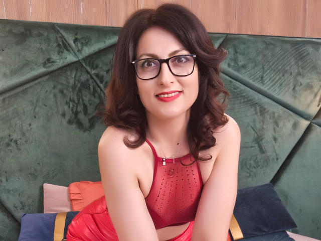 LisaCroftt with curly brown hair, black glasses, red lipstick, red mesh top, and shiny red skirt, smiling, sitting on green velvet couch, showing her small tits, cute.
