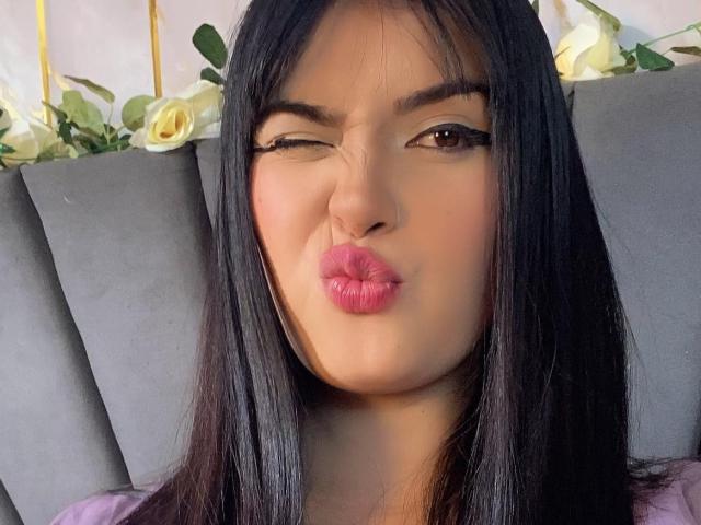 MiaWoodss with long black hair, winking and pouting pink lips, brown eyes, and subtle makeup, sitting on a gray couch with white roses in the background.