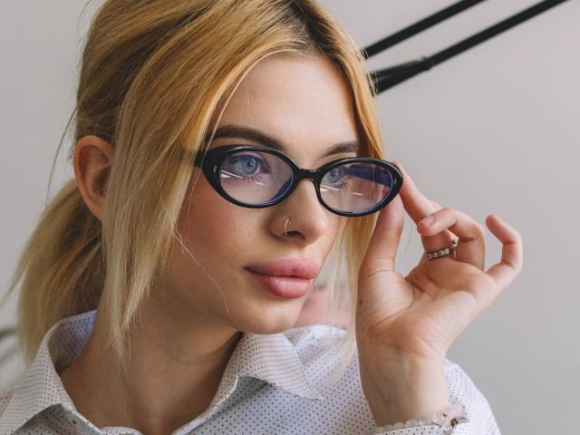AdeleClifford with blonde hair, blue glasses, nose ring, and pink lips, wearing a white patterned shirt, adjusting her glasses with a ring on her right hand.