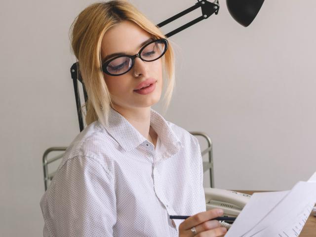 AdeleClifford with blonde hair, black glasses, white polka-dot shirt, holding papers, and a pen, sitting under a desk lamp, looking down with a subtle smile.