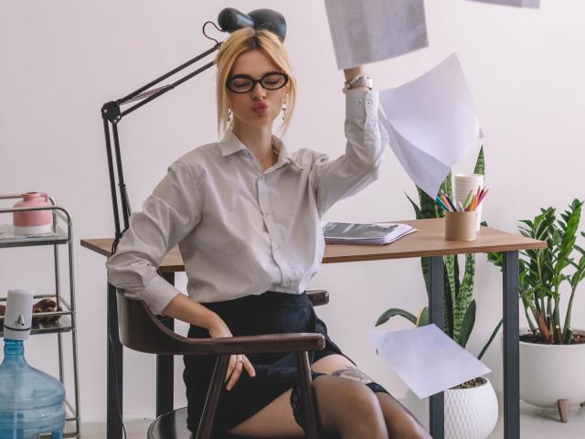 AdeleClifford, blonde, glasses, white shirt, black skirt, garter, sitting, holding papers, desk, lamp, plant, water bottle, office, serious expression, arm raised, papers flying.