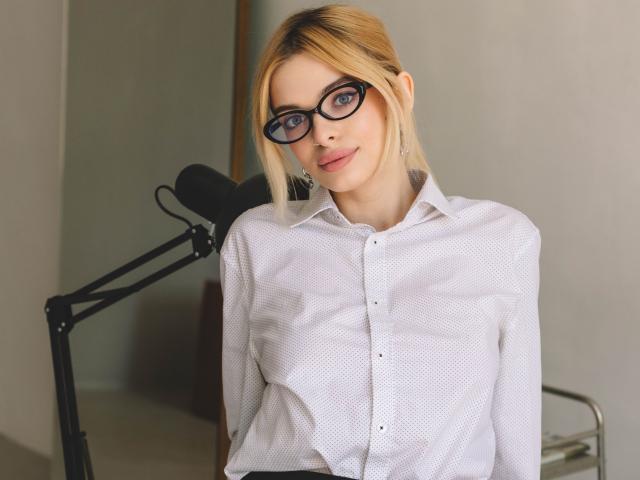 AdeleClifford with blonde hair, glasses, white button-up shirt, and subtle smile, standing in front of a black microphone stand, looking cute and professional.