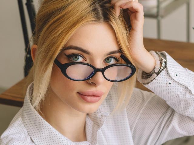 AdeleClifford with blonde hair, green eyes, and black-rimmed glasses, wearing a white polka-dot shirt, touching her hair, with a silver watch on her right wrist.