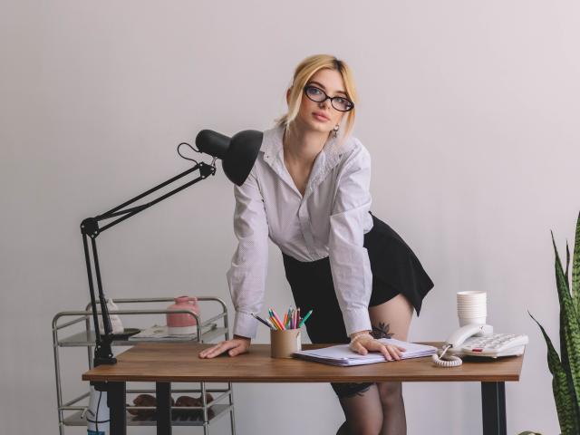 AdeleClifford with blonde hair, glasses, white shirt, black skirt, and thigh tattoo, leaning on desk, pencils, lamp, papers, and phone, with a plant on the right.