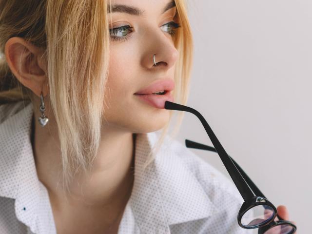 AdeleClifford with blonde hair, nose ring, and heart earrings, biting black glasses, wearing a white checkered shirt, has light pink lipstick, and blue eyes.