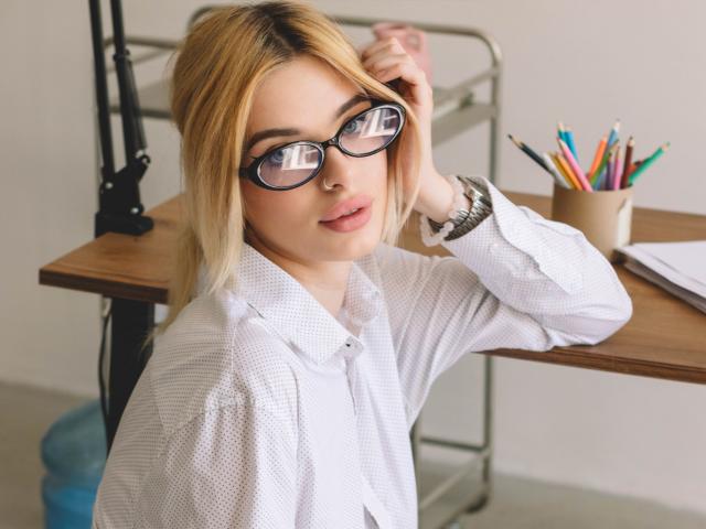 AdeleClifford with blonde hair, glasses, and white shirt, adjusting her hair, sitting at a desk with a pencil holder, looking at the camera with a slight smile.