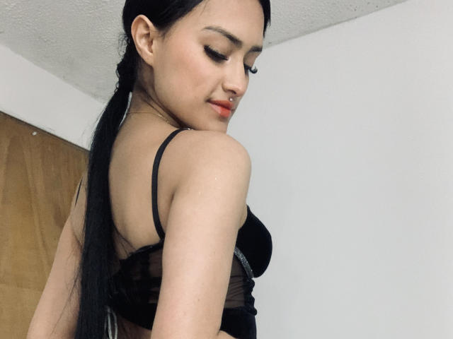 CaassieGlow con cabello negro recogido en una coleta, vistiendo un sostén negro, mostrando sus pequeños senos, sonriendo, mirando hacia abajo, de pie en una habitación con paredes blancas, con una puerta de madera en el fondo..