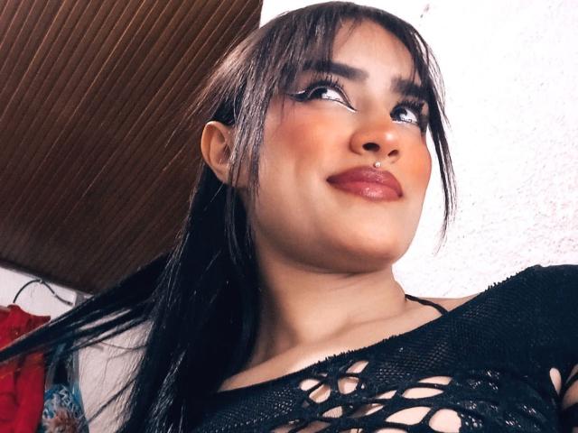 CaassieGlow has long black hair, thick eyeliner, and a nose ring. She's wearing a black, fishnet-style top, showing some skin, with a slight smile and looking up.