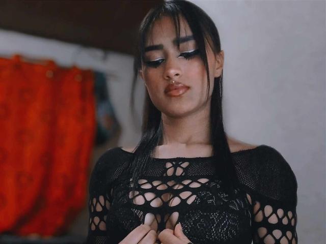 CaassieGlow with black mesh top, showing small tits through open pattern, dark eyeliner, and nose ring, looking down with slightly parted lips. Her black hair is straight and long.