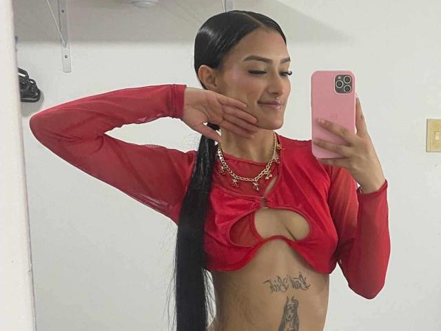Caassie, avec de longs cheveux noirs attachés en queue de cheval, porte un haut rouge transparent avec une découpe, qui révèle ses seins et un tatouage en dessous, et elle prend un selfie..