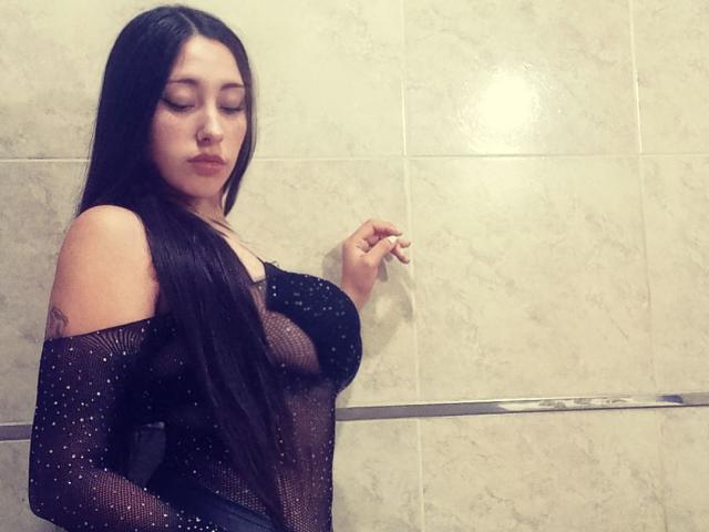 MeganTodaBonita with long black hair, wearing a sheer black top with sparkles, showing her tits, standing against a tiled wall, looking down with a slightly pouty expression.