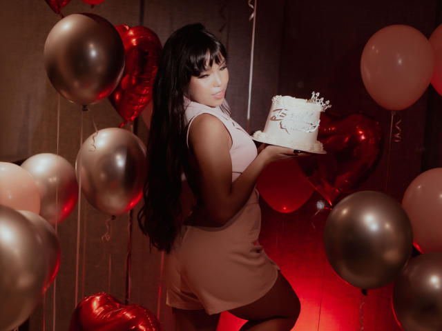 Heyly's holding a white, sparkly cake between her large breasts while surrounded by red and brown balloons, looking back.