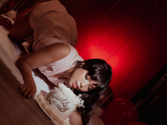 Heyly com cabelo escuro e franja está de quatro, vestindo um top branco, sob intensa iluminação vermelha.