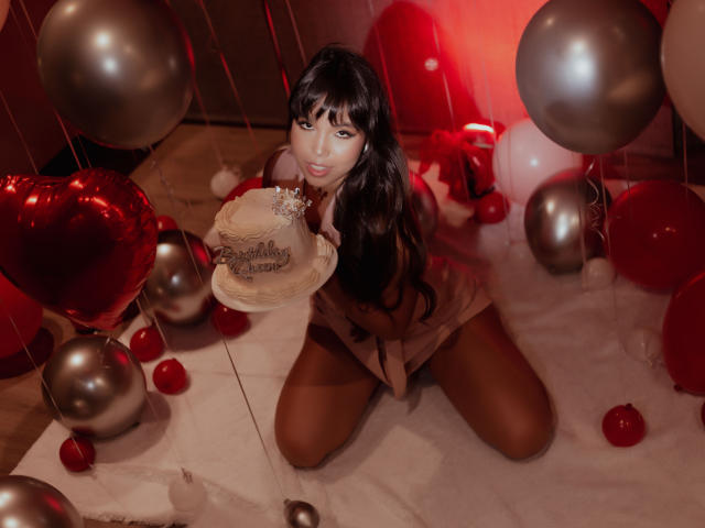 Heyly se arrodilla en una habitación iluminada de rojo rodeada de globos, sosteniendo un sombrero decorado, vistiendo un vestido marrón corto.