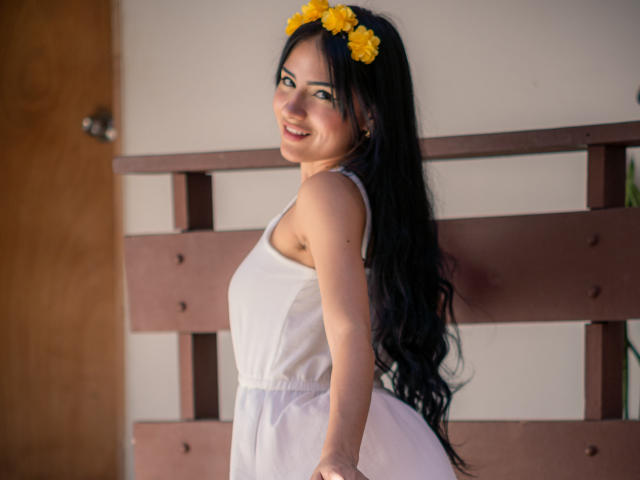 LizzJoy with long black hair, yellow flower headband, white dress, smiling, looking over shoulder, showing her ass, standing in front of brown wooden wall, looking cute.