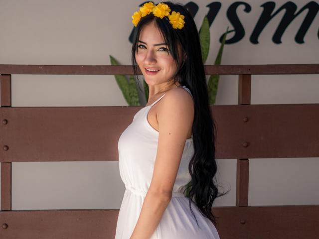 LizzJoy, avec ses longs cheveux noirs, un serre-tête en fleurs jaunes, une robe blanche, souriante, debout, montrant ses fesses, un mur en bois brun, une plante verte, lumière naturelle, mignonne, attirante, peau douce..