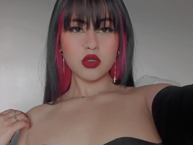 ElisaKim with black and pink hair, red lipstick, nose ring, cross earrings, necklace, and black off-shoulder top, showing her bare tits and holding her shoulder.