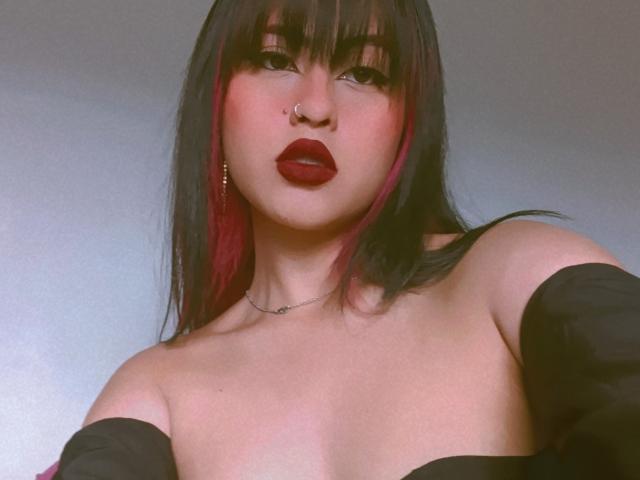 ElisaKim with dark brown hair, red highlights, black off-shoulder top, red lipstick, nose ring, and necklace, showing her bare boobs.