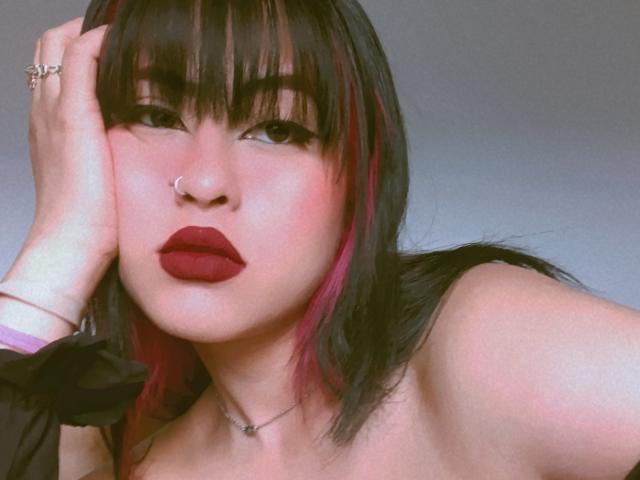ElisaKim has pink and black hair, dark eyeliner, red lipstick, nose ring, and is touching her head with a hand showing a ring. Her tits are partially visible.