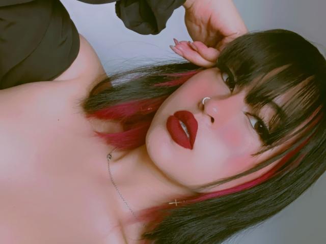 ElisaKim has dark black hair with red tips, red lipstick, a nose ring, and a cross earring, posing with her hand on her head, showing her bare shoulders.