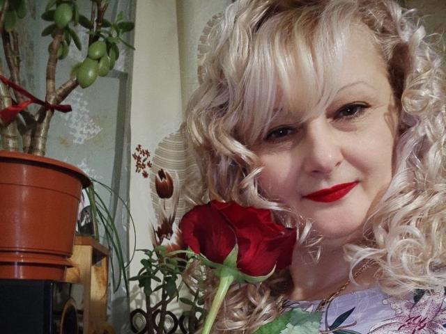 NikolinaBounty with curly blonde hair, red lipstick, and a red rose, smiling at the camera, potted plant to the left, holding rose in front of her tits.