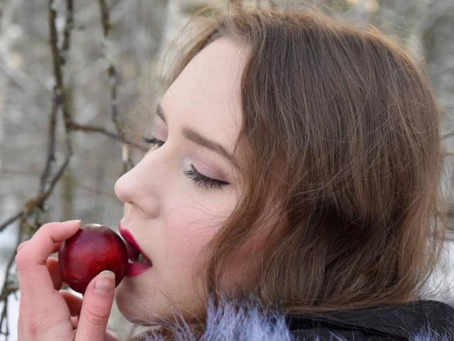 LucyRosen's face, with brown wavy hair, pale skin, and pink eyelids, is eating a red cherry, holding it near her open red-lipped mouth. She's wearing a black jacket.