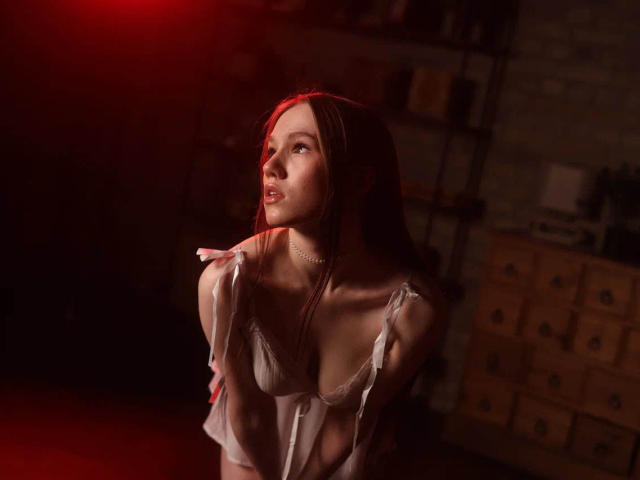 VikkiVibe with red light on her face, looking up, wearing a white, sheer dress, exposing her small tits, holding her shoulder, in a dimly lit room.