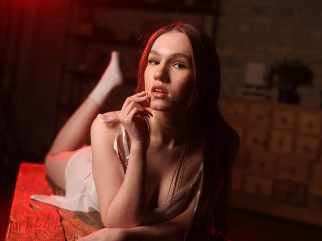 VikkiVibe with long brown hair, lying on a wooden table, wearing white lingerie and socks, biting her finger, red light in the background, showing her tits and soft smile.