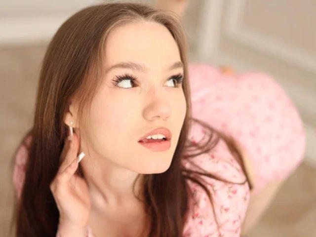VikkiVibe has long brown hair, blue eyes, and is looking to the side with a slight open-mouth expression. She's wearing a pink floral dress, touching her ear.