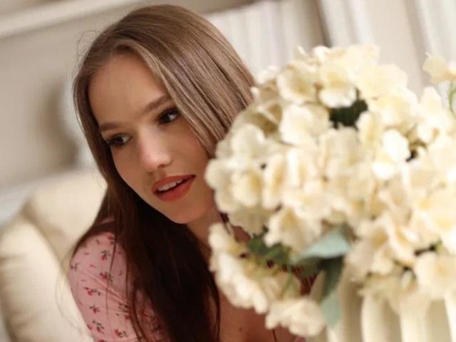 VikkiVibe with long brown hair, pink floral shirt, and white flowers in the foreground, looking slightly to the side with a soft smile. Her tits are partially visible.