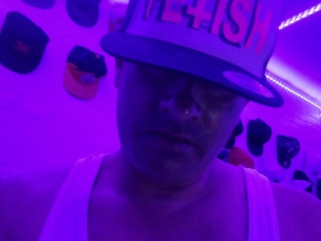 leaveitinsideme-hot wearing a white tank top and a blue and white hat with "METHSH" on it, illuminated by purple neon, showing a serious face.
