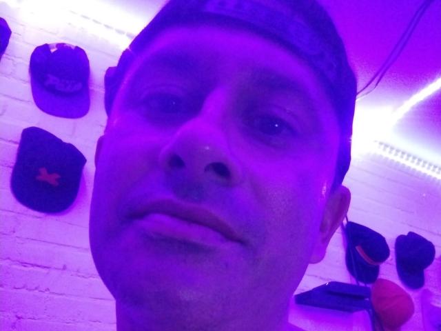 A purple-lit selfie of leaveitinsideme-hot with a serious face, wearing a dark headband, showing a slightly open mouth and subtle nose. Hats are hanging on a white wall.