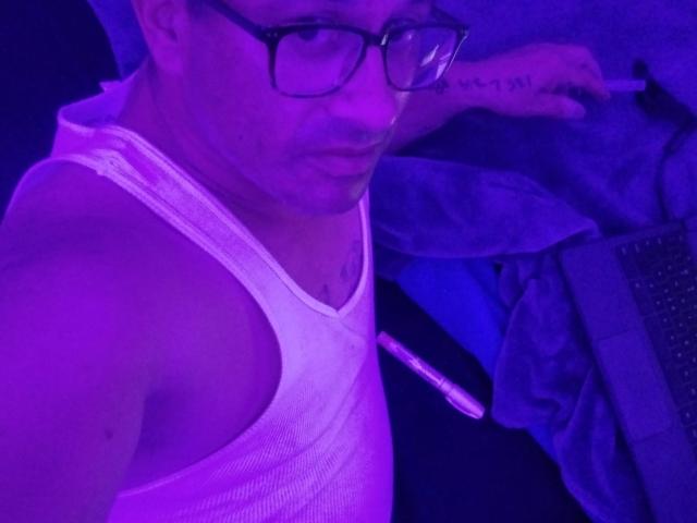Leaveitinsideme-hot wearing a white tank top, purple-lit, with glasses, holding a cigarette, visible tattoo on right arm, slightly pouty expression, and a pen in the left hand.