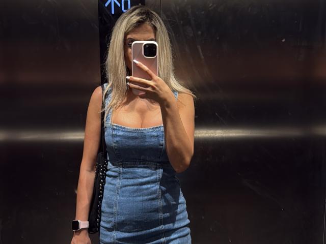 Lellie with blonde hair, wearing a blue denim dress, holding a phone in front of her face, showing her tits, wearing a pink watch, in a mirror selfie.