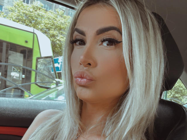Lellie has blonde hair, thick eyebrows, and full lips with a pout. She's wearing long, dramatic eyelashes and looking directly at the camera.