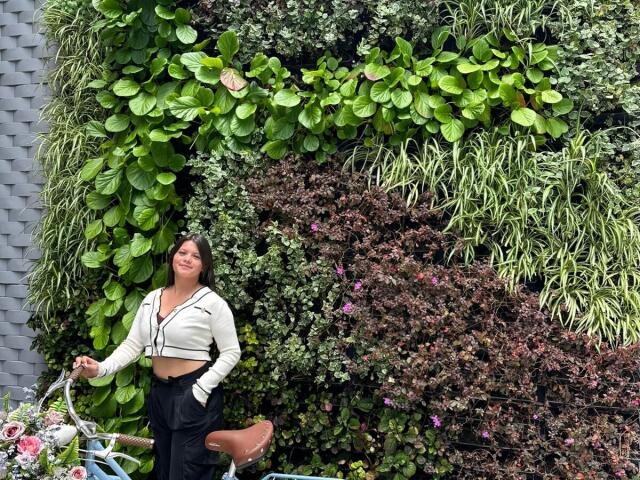 VictoriaWaynes stands in front of a lush green wall, wearing a white crop top and black pants, smiling with brown hair, holding a bike, showing her ass slightly.