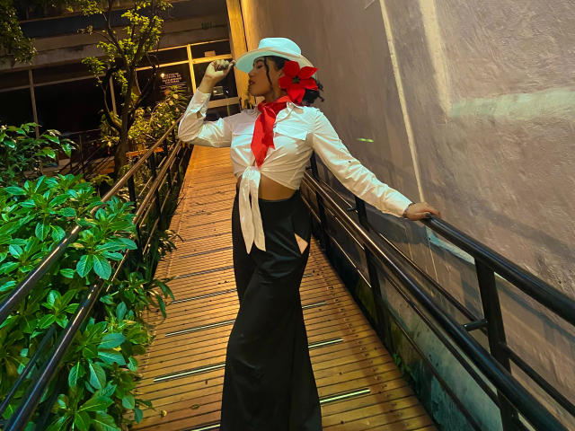 SophieSonyas con grandes senos, vestidas con una camisa blanca atada al frente, falda negra, bufanda roja, sombrero blanco, sosteniendo un teléfono, de pie en un pasillo de madera con barandillas..