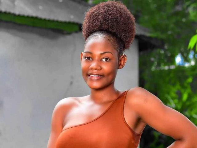 AngeloDivine has dark brown skin, a curly afro, and is wearing a one-shoulder, brown top, smiling with a hand on their hip, showing their small, round tits.