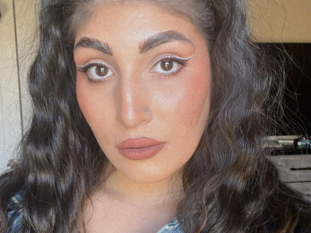 HoneyMoonX has wavy dark brown hair, thick eyebrows, big brown eyes with white eyeliner, a small nose piercing, and full pink lips. Her skin is light and smooth.